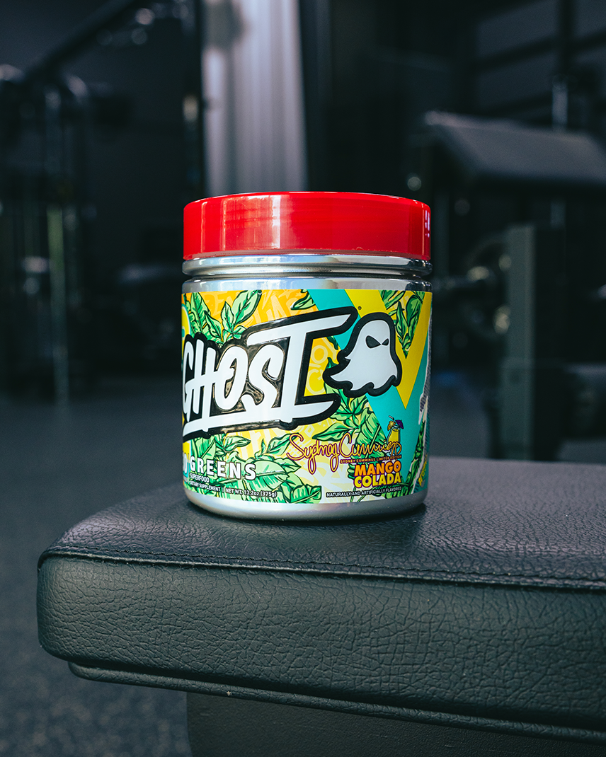A jar of Ghost Labs Energy Enhancers in Lime Eucalyptus flavor, featuring a red lid and a colorful tropical label, resting on a black gym bench with a blurred gym background.